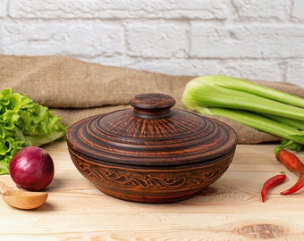 May include: A brown ceramic pot with a lid, featuring a decorative floral design. The pot is sitting on a wooden surface with a red onion, green lettuce, celery stalks, and red chili peppers.