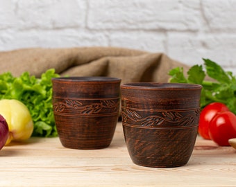 May include: Two rustic, brown ceramic cups with a leaf design. The cups are cylindrical with a dark interior and sit on a light wood surface. Fresh vegetables, including tomatoes and lettuce, are in the background, suggesting a kitchen setting.