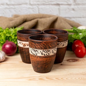 May include: Three brown ceramic mugs with a white floral design. The mugs are on a wooden surface with vegetables in the background.