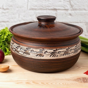 Może przedstawiać: Brązowy ceramiczny garnek do gotowania z pokrywką. Garnek ma ozdobny pas z kwiatowymi wzorami w jaśniejszym kolorze pośrodku. Garnek stoi na drewnianej powierzchni, z warzywami w tle.