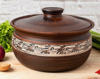 Może przedstawiać: Brązowy ceramiczny garnek do gotowania z pokrywką. Garnek ma ozdobny pas z kwiatowymi wzorami w jaśniejszym kolorze pośrodku. Garnek stoi na drewnianej powierzchni, z warzywami w tle.