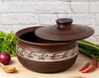 May include: A brown ceramic pot with a lid, featuring a white floral design around the rim. The pot is sitting on a wooden surface with vegetables nearby.