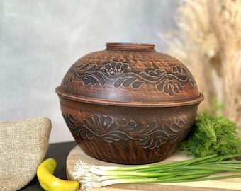 Może przedstawiać: Brązowy ceramiczny garnek do gotowania z pokrywką, zdobiony kwiatowymi rzeźbami. Garnek stoi na drewnianej desce do krojenia z zieloną cebulą, koperkiem i żółtą papryką. Po lewej stronie worek jutowy.