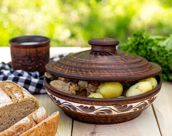 Könnte beinhalten: Eine braune Keramikschüssel mit Deckel, gefüllt mit gekochten Kartoffeln und Fleisch, steht auf einem Holztisch. Geschnittenes Brot, eine Tasse Milch und Grünzeug sind ebenfalls vorhanden. Die Schüssel hat ein dekoratives weißes und braunes Muster.