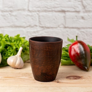 Peut inclure: Une tasse en céramique brune avec une surface texturée est posée sur une table en bois. Il y a des feuilles de laitue verte, des bulbes d'ail et un poivron rouge en arrière-plan.