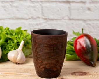 May include: A brown ceramic mug with a textured surface sits on a wooden table.  There are green lettuce leaves, garlic bulbs, and a red bell pepper in the background.