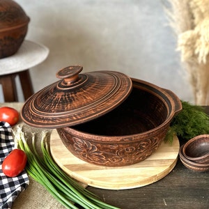 Puede incluir: Una olla de cerámica marrón con tapa, con un diseño floral. La olla está sobre una tabla de cortar de madera, rodeada de verduras frescas. La olla tiene aproximadamente 25 cm de diámetro.