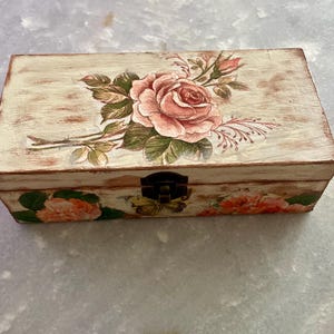 Op de afbeelding: Een rechthoekige houten kist met een verweerde crème afwerking. De deksel is versierd met een grote roze roos en groene bladeren. De voorkant van de kist heeft een kleiner rozenontwerp. Een metalen sluiting zekert de kist.