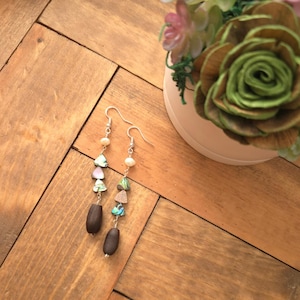 May include: A pair of dangle earrings with silver hooks. Each earring features a sequence of small, colorful, triangular and oval-shaped beads, including abalone shell, pink, and brown wooden beads. The earrings are displayed on a wooden surface.