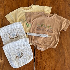 Puede incluir: Un conjunto de ropa y accesorios para bebés en tonos neutros. Incluye una camiseta amarilla, un body marrón con bordados de patos y el nombre "Beau Allan", dos baberos blancos con un diseño de cornamenta y un clip para chupete.