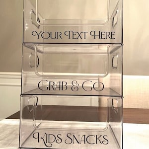 Puede incluir: Tres contenedores de almacenamiento apilables de plástico transparente con etiquetas negras. El contenedor superior dice "YOUR TEXT HERE", el contenedor del medio dice "GRAB & GO" y el contenedor inferior dice "KIDS SNACKS".