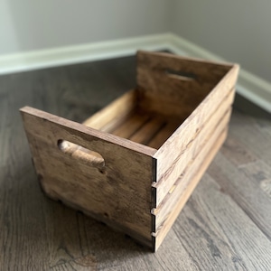 May include: A rustic wooden crate with a distressed brown finish. The crate has two cut-out handles on the side and is made of natural wood.