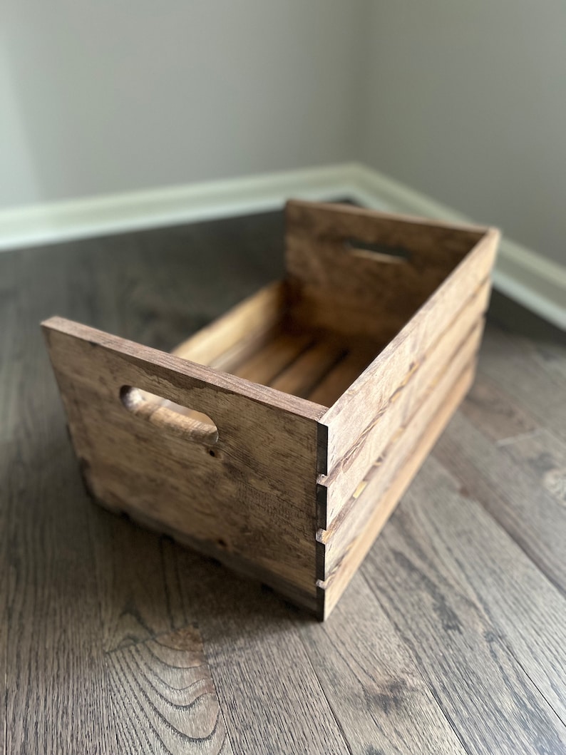 May include: A rustic wooden crate with a distressed brown finish. The crate has two cut-out handles on the side and is made of natural wood.
