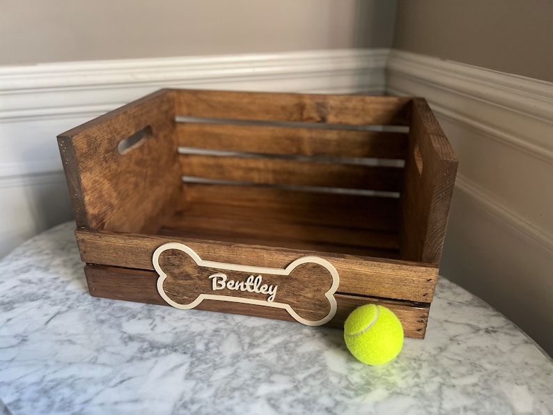May include: A wooden dog crate with a bone-shaped name tag that says "Bentley". A yellow tennis ball is in front of the crate.