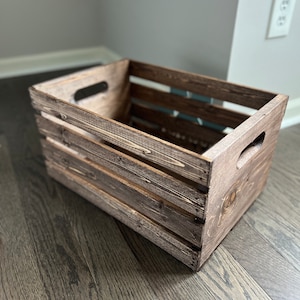 May include: A rustic wooden crate with a dark brown stain. The crate has a rectangular shape with a slatted design and two handles on the sides.