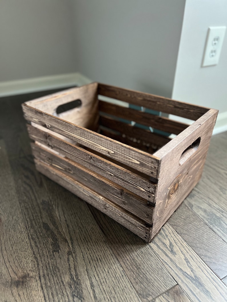 May include: A rustic wooden crate with a dark brown stain. The crate has a rectangular shape with a slatted design and two handles on the sides.