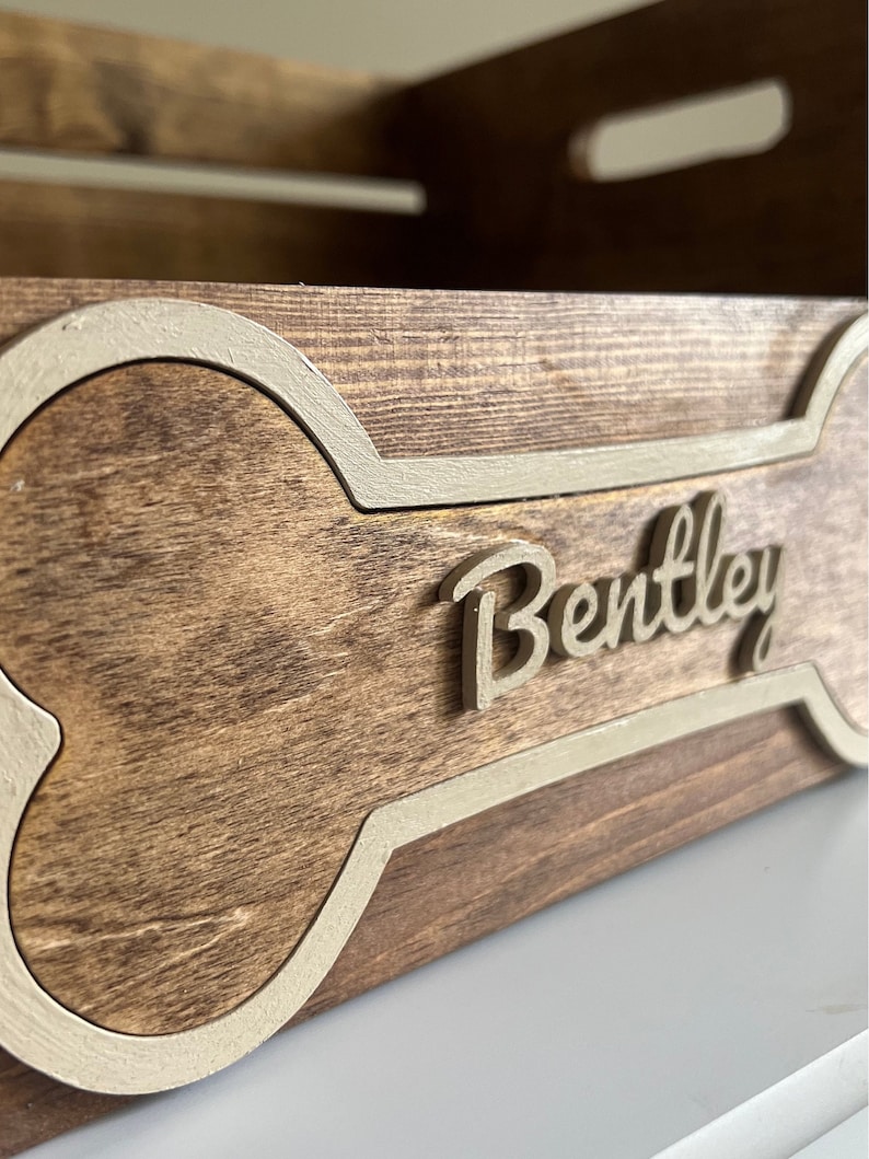 May include: A wooden dog bed with a bone-shaped cutout and the name "Bentley" engraved on the front. The bed is stained a dark brown color and has a white painted border around the bone shape.