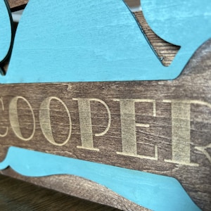 May include: A wooden dog bone sign with a light blue painted outline. The name "COOPER" is engraved in gold lettering on the bone.