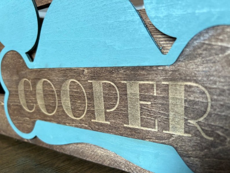 May include: A wooden dog bone sign with a light blue painted outline. The name "COOPER" is engraved in gold lettering on the bone.