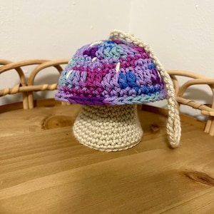 May include: A crocheted mushroom ornament with a purple, blue, and white patterned cap and a beige stem. The ornament has a loop for hanging.