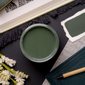 May include: A small jar of dark green paint with a white lid. The paint is on a black surface with a book and a piece of paper with a sample of the paint.