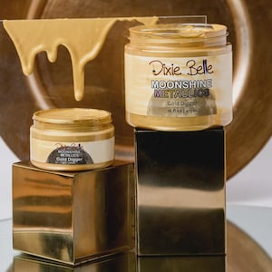 May include: Two jars of Dixie Belle Moonshine Metallics paint in the color Gold Digger. The larger jar is 16 fl oz and the smaller jar is 4 fl oz. The paint is a metallic gold color, and a sample of the paint is shown dripping from a clear sheet.