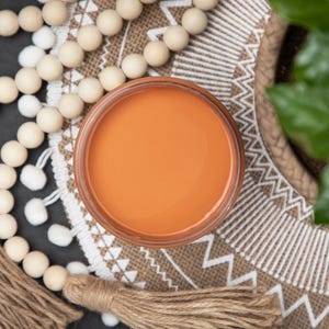 May include: A round glass jar filled with smooth, vibrant orange liquid, positioned centrally. The jar is surrounded by a long strand of natural wooden beads featuring two tan jute tassels at the ends, resting on a circular woven mat.
