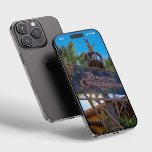 May include: A black iPhone with a screen displaying a photo of the Pirates of the Caribbean ride at a theme park. The screen shows a sign with the words "Pirates of the Caribbean" and a pirate ship in the background.