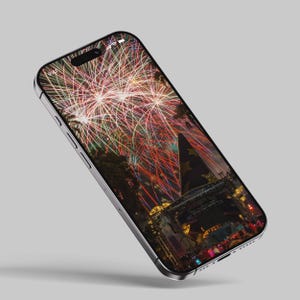 May include: A smartphone displaying a photo of fireworks exploding over a crowd of people gathered in front of a stage. The fireworks are colorful and create a vibrant display in the night sky.