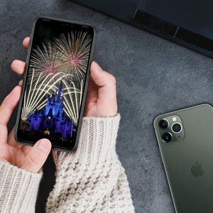 May include: A person holding a black smartphone with a photo of fireworks over a castle on the screen. A green smartphone with a triple lens camera is lying on a gray surface.