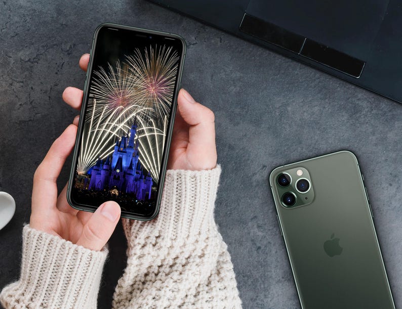 May include: A person holding a black smartphone with a photo of fireworks over a castle on the screen. A green smartphone with a triple lens camera is lying on a gray surface.