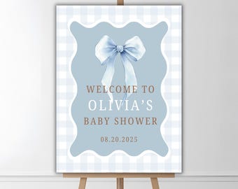 May include: A baby shower welcome sign on an easel. The sign features a light blue background with a white bow illustration. The text reads "Welcome to Olivia's Baby Shower" and the date "08.20.2025". The background has a light blue and white checkered pattern.