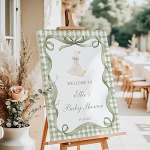 Può includere: Un cartello di benvenuto per il baby shower su un cavalletto. Il cartello presenta un'illustrazione ad acquerello di un'oca con un fiocco, circondata da un bordo e un nastro a quadretti verdi. Il testo recita "Benvenuti al baby shower di Ellie" e la data.