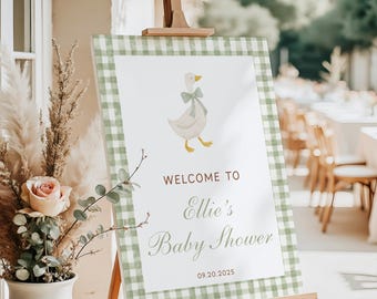 May include: A baby shower welcome sign on a wooden easel. The sign features a white background with a green gingham border. The sign has a cartoon goose with a bow and the text "Welcome to Ellie's Baby Shower" and the date 09.20.2025.