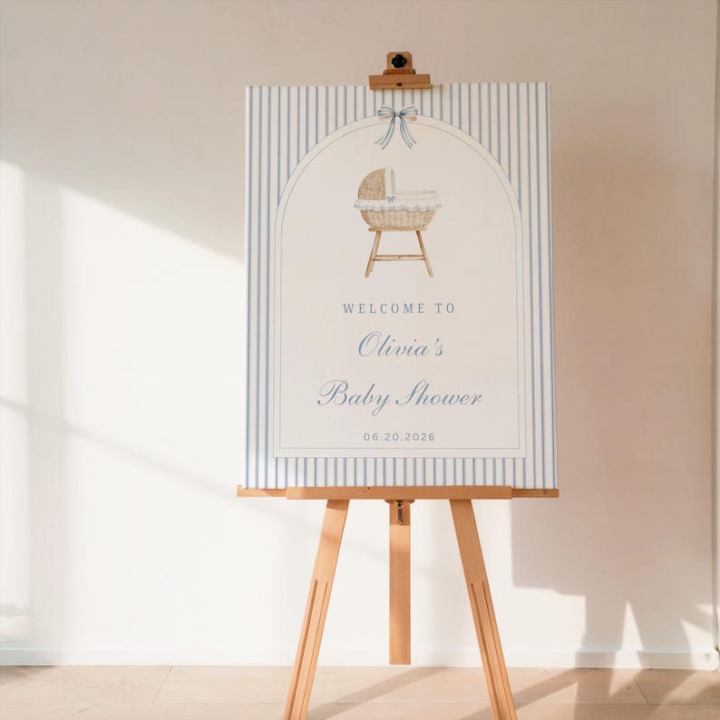 May include: A baby shower sign on a wooden easel. The sign features a blue and white striped background with an arched design. The design includes a baby bassinet illustration and the text "Welcome to Olivia's Baby Shower" with the date 06.20.2026.