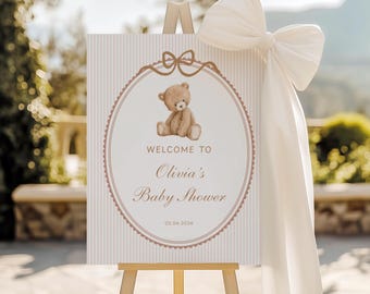 May include: A baby shower welcome sign on a wooden easel. The sign features a teddy bear illustration inside a decorative oval frame. The text reads "WELCOME TO Olivia's Baby Shower 02.06.2026". A large white bow adorns the top.
