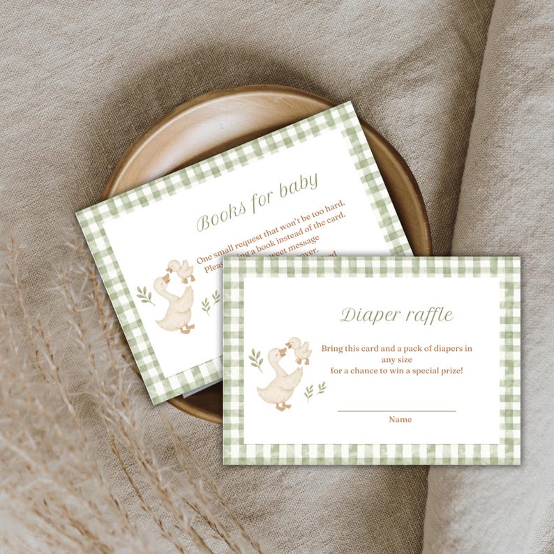 May include: Baby shower invitation cards with a green gingham border. The cards feature illustrations of geese and the text "Books for baby" and "Diaper raffle". The cards are on a wooden plate.