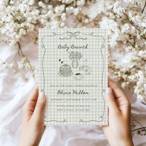 May include: A baby shower invitation with a light green gingham background and a decorative border. The invitation features the text "Baby Brunch" and illustrations of breakfast items. The invitation is held by two hands.