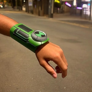 May include: A green and black wristwatch with a large, circular dial featuring a green alien symbol. The watch has a textured, rubber-like band.