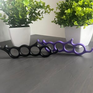 May include: Two knuckle-duster style rings, one black and one purple, are displayed on a grey surface. Each ring has four finger holes and a curved handle. Two small potted plants with green foliage are in the background.