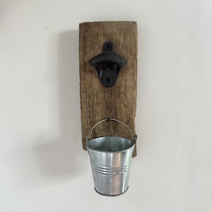 May include: A rustic wall-mounted bottle opener with a galvanized metal bucket. The opener is made of black metal and is attached to a weathered wooden plank. The bucket hangs below, ready to catch bottle caps. The opener reads "OPEN HERE".
