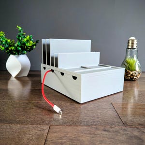 May include: White plastic charging station with multiple slots for devices. A red charging cable is plugged into the station and extends to the floor.