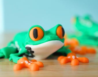Op de afbeelding: Een groene kikkerfiguur met oranje voeten en grote zwarte ogen. De kikker ligt op een houten oppervlak.