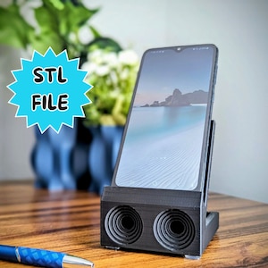 Puede incluir: Un soporte de teléfono negro con dos altavoces en la parte delantera. El soporte sostiene un teléfono inteligente con una imagen azul y blanca en la pantalla. El texto "STL FILE" está en una estrella azul.