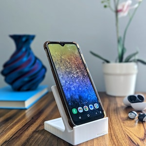 May include: A white phone stand with a black smartphone in a vertical position. The phone screen displays a variety of colourful icons. The stand is on a wooden surface.