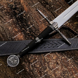 Könnte beinhalten: Ein schwarzer Ledergürtel mit einem dekorativen Muster und ein silbernes Schwert mit einem gedrehten Griff und einem schwarzen Griff. Das Schwert hat einen silbernen Knauf mit einem dekorativen Design.