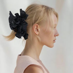 Peut inclure: Une pince à cheveux en tissu noir avec un design plissé. La pince a la forme d'une fleur.