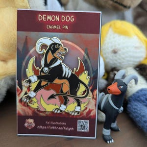 May include: A black, orange, and white enamel pin of a demon dog with flames, next to a small figurine of the same character. The pin is on a card that reads "DEMON DOG ENAMEL PIN". The background is blurred with plush toys.