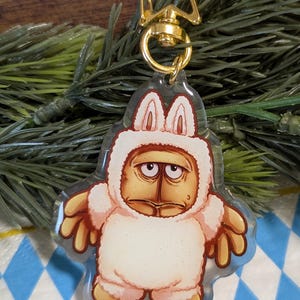 May include: A clear acrylic keychain featuring a cartoon character in a white bunny suit with a grumpy expression. The keychain has a gold-colored star-shaped clasp and a gold-colored swivel clip.
