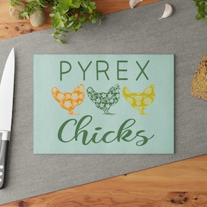 May include: A rectangular Pyrex cutting board with the words "PYREX Chicks" in green. The board features three floral-patterned chickens in orange, green, and yellow. The board is on a gray surface with a knife and greenery in the background.